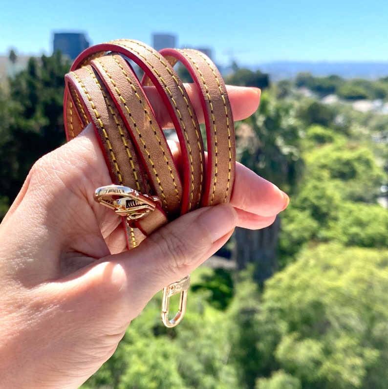 May include: A brown leather strap with red stitching and a gold metal clasp. The strap is coiled and held in a hand.