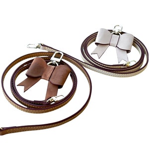 May include: Two leather straps with brown and beige stitching. Each strap has a metal loop and a leather bow. The bow on the left strap is brown and the bow on the right strap is beige.