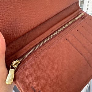 May include: Open brown leather wallet with a gold zipper and a gold zipper pull with the Louis Vuitton logo. The wallet has multiple card slots and a textured surface. The interior is a lighter shade of brown.