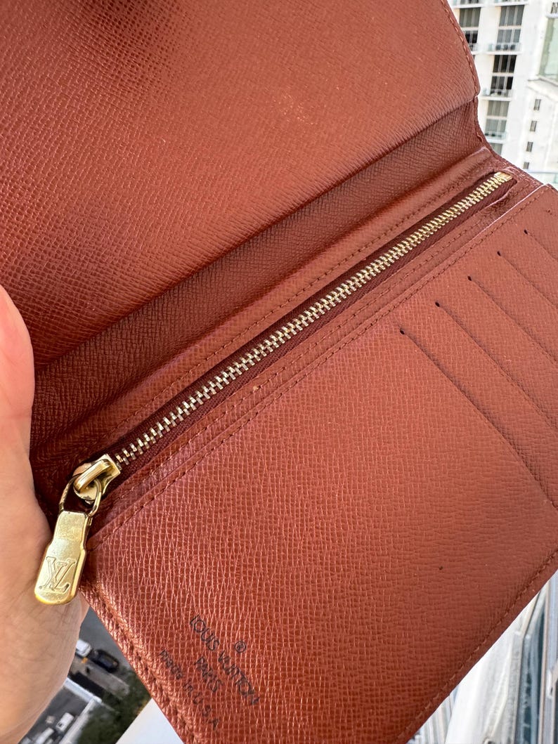 May include: Open brown leather wallet with a gold zipper and a gold zipper pull with the Louis Vuitton logo. The wallet has multiple card slots and a textured surface. The interior is a lighter shade of brown.