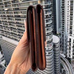 May include: A brown leather wallet with a bi-fold design. The wallet is held vertically, showcasing its interior and exterior. The exterior is a darker brown, while the interior is a lighter shade. The background features a cityscape with tall buildings.
