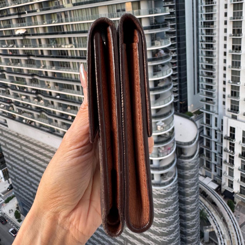 May include: A brown leather wallet with a bi-fold design. The wallet is held vertically, showcasing its interior and exterior. The exterior is a darker brown, while the interior is a lighter shade. The background features a cityscape with tall buildings.