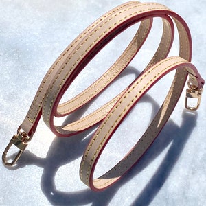 May include: A beige leather strap with red stitching and gold hardware. The strap is folded in a loop and has a hook on each end.