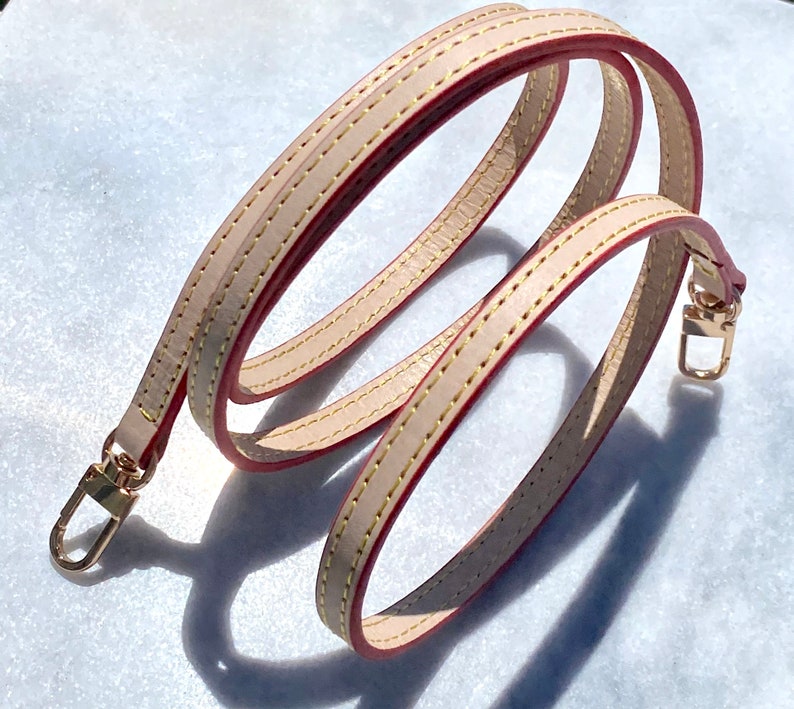 May include: A beige leather strap with red stitching and gold hardware. The strap is folded in a loop and has a hook on each end.
