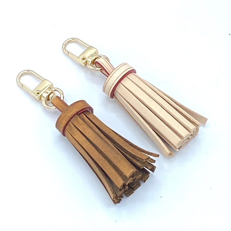 May include: Two leather keychains with tassels. One is brown with a red stripe and the other is beige with a red stripe.