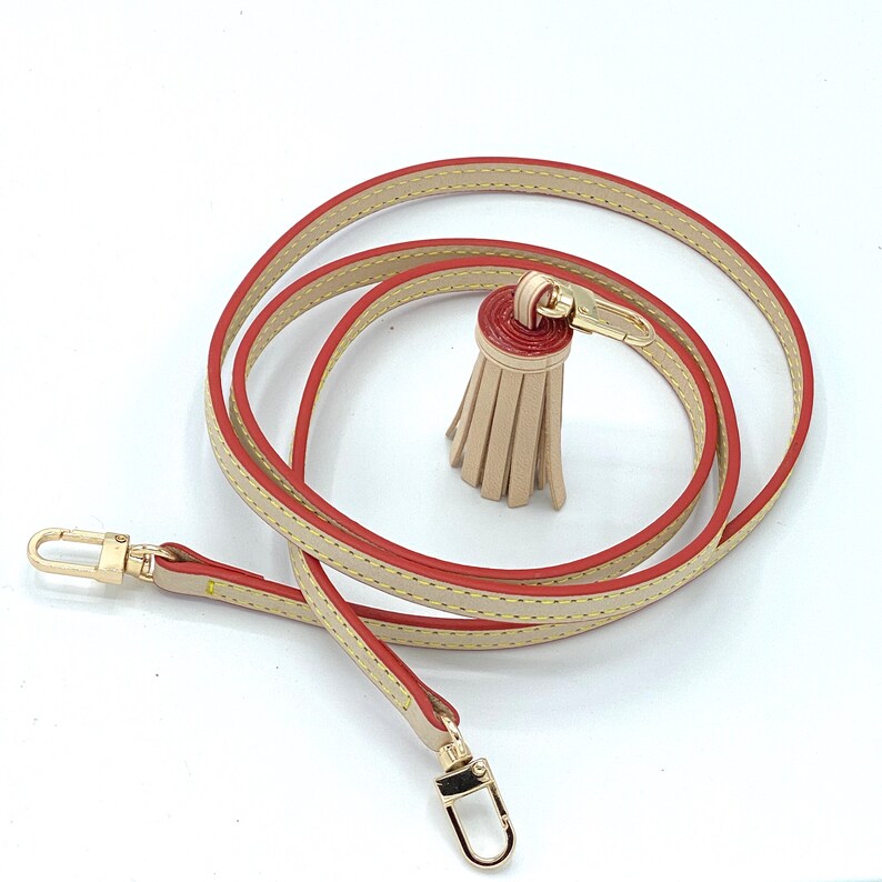 May include: A beige leather strap with a red and beige tassel. The strap has gold hardware and is approximately 50 inches long.