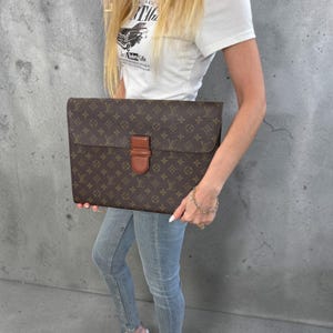 May include: A brown document case featuring the iconic Louis Vuitton monogram pattern in gold and dark brown. The rectangular bag has a fold-over flap secured by a single tan leather tab closure centered on the front. The material appears to be coated canvas with leather trim.