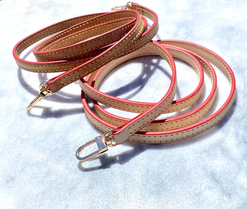 May include: A beige leather strap with a red edge and gold metal clasps. The strap is folded and coiled on a white surface.