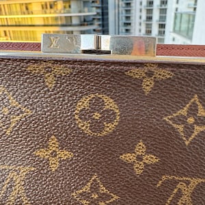 May include: Close-up of a brown handbag with a gold monogram pattern. The bag features a silver clasp with the brand's logo. The background shows a cityscape with tall buildings.