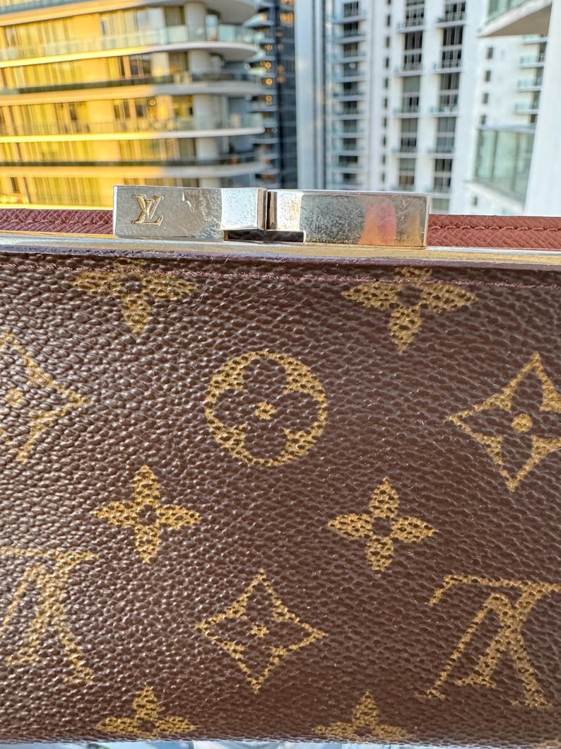 May include: Close-up of a brown handbag with a gold monogram pattern. The bag features a silver clasp with the brand's logo. The background shows a cityscape with tall buildings.