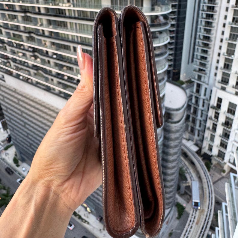 May include: A brown leather wallet with a flap closure. The wallet is held up against a backdrop of a city skyline. The wallet appears to be made of high-quality leather and has a classic design. The wallet is a bi-fold style.
