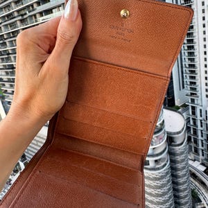 May include: Open brown leather wallet with multiple card slots and a snap closure. The interior is a lighter shade of brown. The brand name "LOUIS VUITTON PARIS made in France" is visible on the inside.