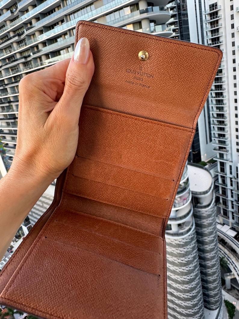 May include: Open brown leather wallet with multiple card slots and a snap closure. The interior is a lighter shade of brown. The brand name "LOUIS VUITTON PARIS made in France" is visible on the inside.