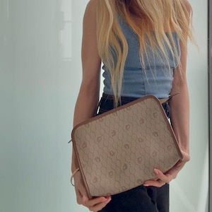 May include: A beige and brown handbag with a repeating pattern. The bag has a rectangular shape with rounded corners and a brown border. The bag is held in front of a person wearing a blue top and black pants.