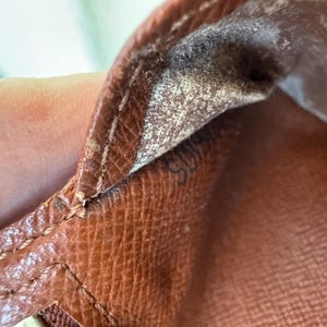 May include: Close-up of a brown leather item, possibly a wallet or handbag, showing detailed stitching and the texture of the material. The interior lining is visible, with a faded pattern and a small, barely visible marking.