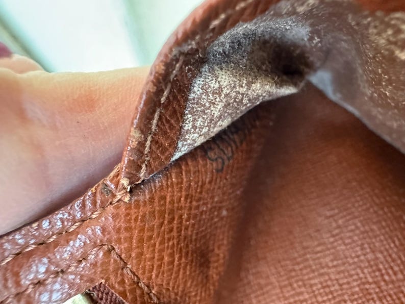 May include: Close-up of a brown leather item, possibly a wallet or handbag, showing detailed stitching and the texture of the material. The interior lining is visible, with a faded pattern and a small, barely visible marking.