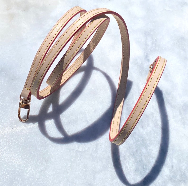 May include: A beige leather strap with red stitching and a gold metal clasp. The strap is coiled and appears to be a replacement strap for a handbag.