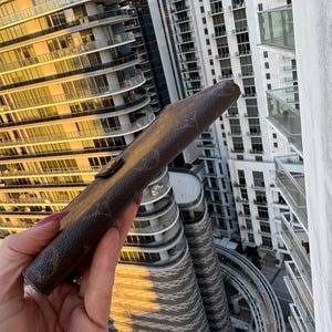 May include: A brown leather case with a monogram pattern, held against a backdrop of high-rise buildings. The case has a flap closure with a gold-colored button. The case appears to be a wallet or a similar accessory.
