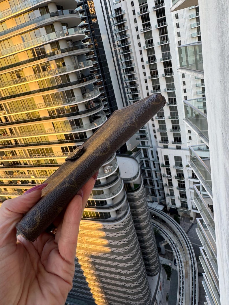 May include: A brown leather case with a monogram pattern, held against a backdrop of high-rise buildings. The case has a flap closure with a gold-colored button. The case appears to be a wallet or a similar accessory.