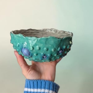 以下が含まれることがあります： 手作りのセラミックボウル。テクスチャ加工されたターコイズブルーの外側とグレーの内側。 ボウルは、隆起した丸みを帯びたディテールといくつかの紫色のアクセントが特徴です。 リムは波状の有機的な形状をしています。