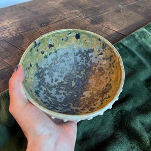 May include: A round ceramic bowl featuring a reactive glaze with a mottled pattern of olive green, pale blue, and dark charcoal gray speckles. The exterior rim of the bowl displays a series of small, evenly spaced raised bumps or studs.