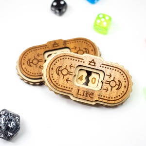 May include: Two wooden life counters with engraved designs and the word "LIFE". The counters display the number 40. Various dice in black, green, and blue are scattered around the counters on a white surface. The counters are for tabletop games.