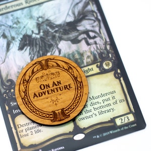 May include: A wooden token with the text "ON AN ADVENTURE" engraved on it. The token is round and has a decorative border. It is sitting on top of a Magic: The Gathering card.