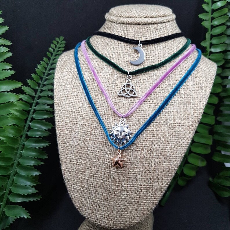 May include: Four velvet choker necklaces with different charms. The charms include a crescent moon, a triquetra, a sun, and a star. The necklaces are displayed on a brown fabric mannequin.