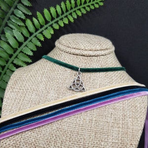 May include: A green velvet choker necklace with a silver Celtic knot pendant. The necklace is displayed on a fabric-covered mannequin with multiple velvet ribbons in various colors.