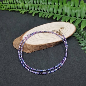 Op de afbeelding: Een delicate ketting met twee rijen van kleine paarse, blauwe en zilveren kralen. De ketting heeft een gouden sluiting.