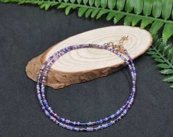 May include: A delicate double-strand necklace made with tiny purple, blue, and silver beads. The necklace has a gold clasp.