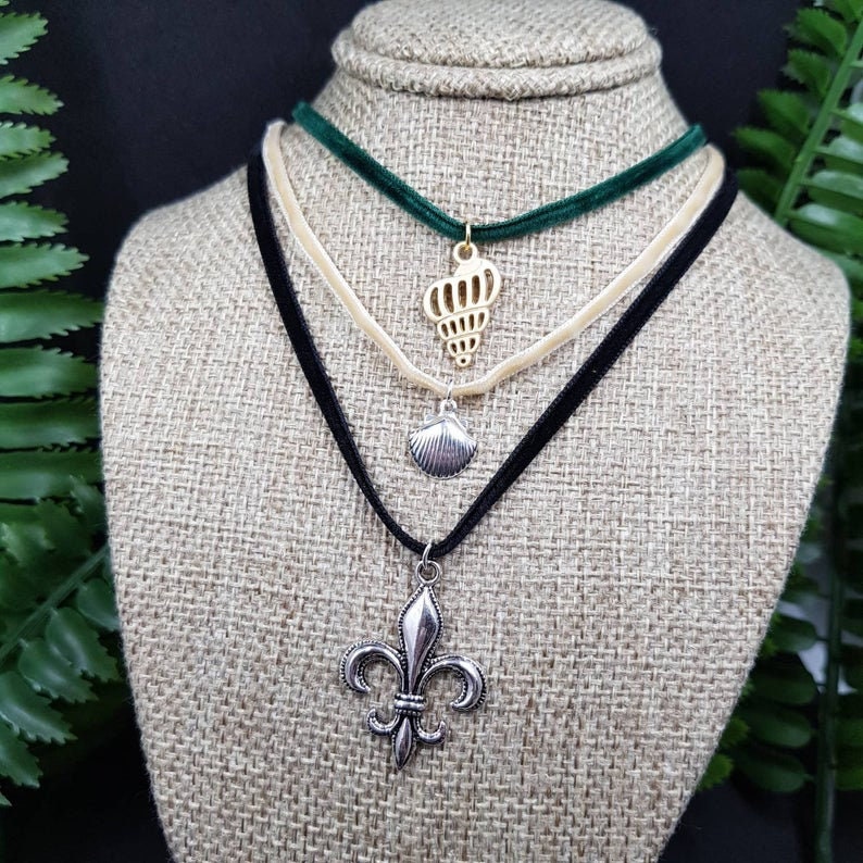 May include: Three layered velvet choker necklaces with different charms. The top layer is green velvet with a gold crown charm. The middle layer is cream velvet with a gold seashell charm. The bottom layer is black velvet with a silver fleur-de-lis charm.