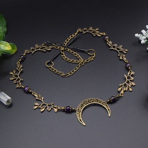 Könnte beinhalten: Ein antikgoldenes Haaraccessoire mit einem Halbmond- und Blätterdesign. Das Haarteil ist mit kleinen lila Perlen verziert und enthält Haarnadeln und eine Kette. Das Design erinnert an Themen aus dem Bereich Himmel und Natur.