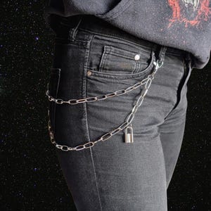 Könnte beinhalten: Eine silberne Kette mit einem Vorhängeschloss, die um die Taille einer Person getragen wird, die eine schwarze Jeans und ein schwarzes Kapuzenpullover trägt.