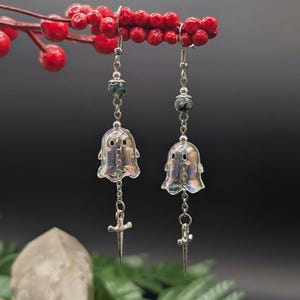 Puede incluir: Un par de pendientes colgantes con dijes iridiscentes en forma de fantasma, peque&ntilde;as cuentas moteadas de color verde oscuro y colgantes de espada plateados que cuelgan de cadenas de metal. Los pendientes cuelgan de ganchos plateados.