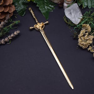 Puede incluir: Un accesorio para el cabello de metal dorado con forma de espada en miniatura. La pieza presenta una hoja larga y puntiaguda, una guarda detallada con un acento triangular central y un mango delgado con un pequeño lazo en el extremo.