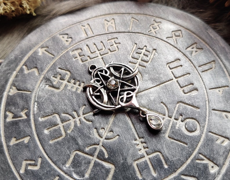 May include: A silver-colored pendant featuring a pentagram design with a small, clear gemstone in the center. The pendant hangs from a teardrop-shaped gemstone. The pendant rests on a gray, circular stone etched with symbols.