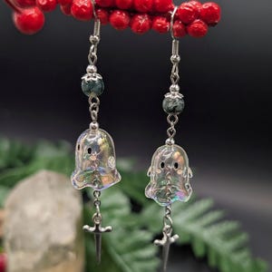 Puede incluir: Un par de pendientes colgantes con dijes de fantasmas iridiscentes y transl&uacute;cidos con ojos negros peque&ntilde;os y mejillas rosadas. Cada fantasma cuelga de una cadena plateada con una cuenta jaspeada verde oscuro y un peque&ntilde;o dije de espada.