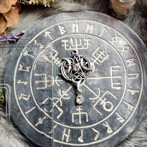 May include: A silver pendant featuring a pentagram with a central crystal and a teardrop-shaped crystal below. The pendant is approximately 1.5 inches in length and is displayed on a gray, circular stone with engraved symbols. The background includes natural elements.