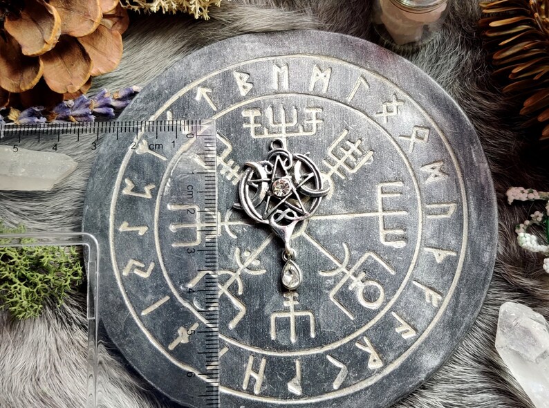 May include: A silver pendant featuring a pentagram with a central crystal and a teardrop-shaped crystal below. The pendant is approximately 1.5 inches in length and is displayed on a gray, circular stone with engraved symbols. The background includes natural elements.