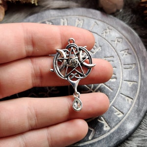 May include: A silver-toned pendant featuring a pentagram with a central crystal and crescent moons. The pendant has a teardrop-shaped crystal dangling below. The design incorporates Celtic knotwork and is held in a hand.