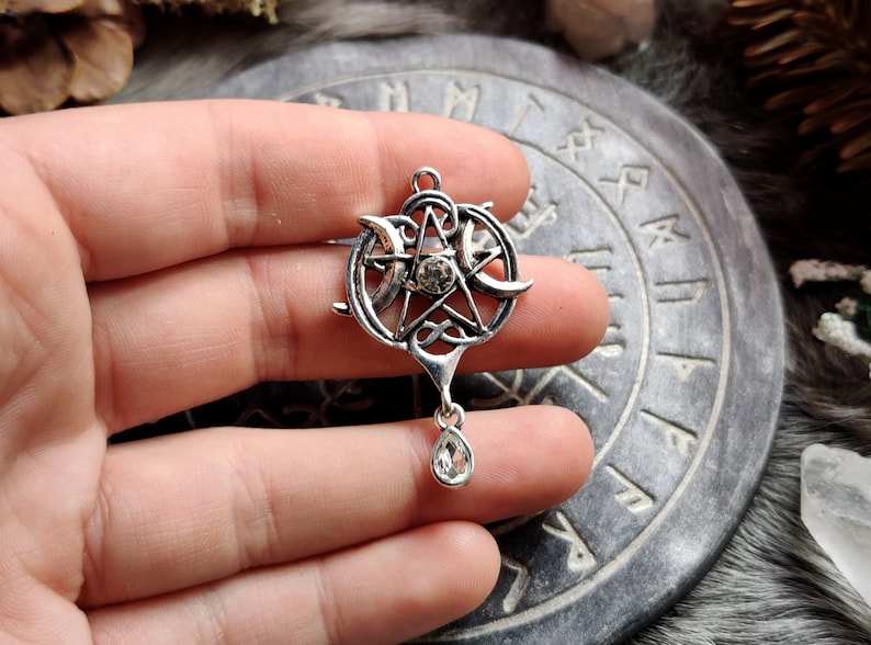 May include: A silver-toned pendant featuring a pentagram with a central crystal and crescent moons. The pendant has a teardrop-shaped crystal dangling below. The design incorporates Celtic knotwork and is held in a hand.