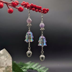 Puede incluir: Un par de pendientes colgantes con gancho plateado, una cuenta redonda de color violeta claro, un dije iridiscente con forma de fantasma y un pequeño dije de bellota plateado en la parte inferior. Los pendientes cuelgan de una rama de bayas rojas.
