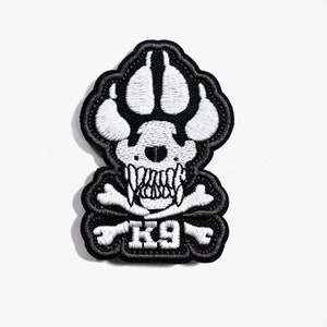 May include: Embroidered patch with a dog paw print skull design and crossbones. The patch is black with white stitching, and the text "K9" is at the bottom. The patch has a black border.