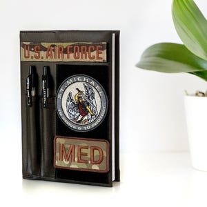 May include: A dark brown leather notebook with a pen holder and patches. The patches read "U.S. AIR FORCE", "ST. MICHAEL", and "MED". Two black pens are in the pen holder. The notebook is on a white surface.