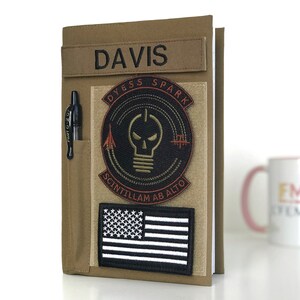May include: Tan notebook cover with a pen holder and patches. The patches include the name "DAVIS", a circular design with a skull and lightbulb, and a black and white American flag. A black pen is in the holder.