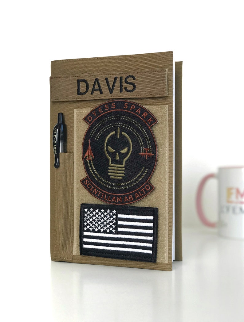 May include: Tan notebook cover with a pen holder and patches. The patches include the name "DAVIS", a circular design with a skull and lightbulb, and a black and white American flag. A black pen is in the holder.