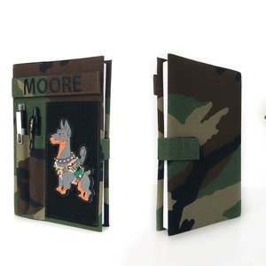 May include: Camouflage-patterned notebook cover with the name "MOORE" embroidered in black. Features a black patch with a cartoon dog design, pen holders, and a green strap closure. The notebook is open, revealing white pages.