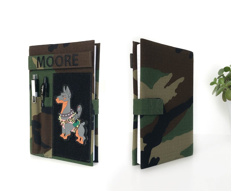 May include: Camouflage-patterned notebook cover with the name "MOORE" embroidered in black. Features a black patch with a cartoon dog design, pen holders, and a green strap closure. The notebook is open, revealing white pages.