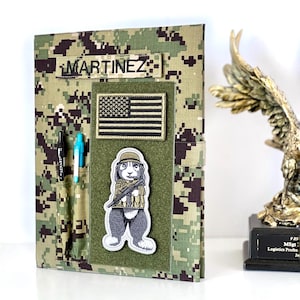 May include: A digital camouflage patterned notebook cover featuring a name patch that reads MARTINEZ, a United States flag patch, and an embroidered patch of a rabbit in military gear. Two pen slots hold a black pen and a light blue pen.